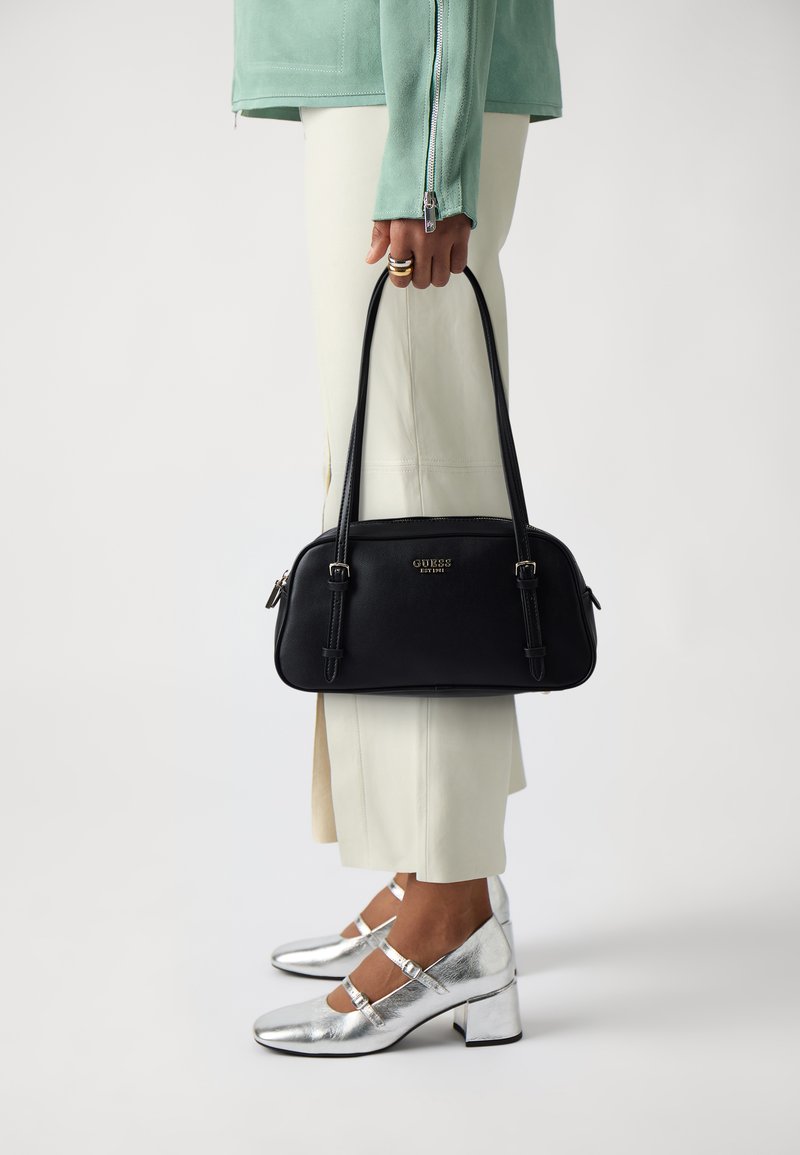 Bolso negro con dobles asas, logo dorado, diseño elegante, textura suave, combinado con pantalones crema y tacones plateados metálicos.