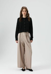 Zwarte gebreide cardigan met een cropped ontwerp en knoopsluiting. Gecombineerd met beige, wijde, high-waisted broek en zwarte instapschoenen.