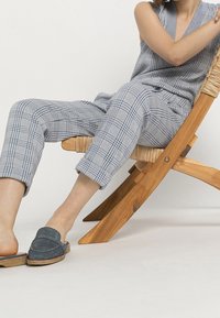 Gray and white plaid sleeveless jumpsuit with a relaxed fit, paired with navy slip-on shoes, seated on a wooden chair with woven details.