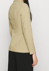 Blazer beige ajusté avec une texture lisse, coupe slim, col cranté et manches longues, associé à un pantalon noir. Design simple et épuré.