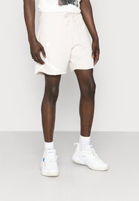 Shorts sportivi beige in cotone, con vita elasticizzata e cordino, e una grafica bianca del logo sulla gamba sinistra.