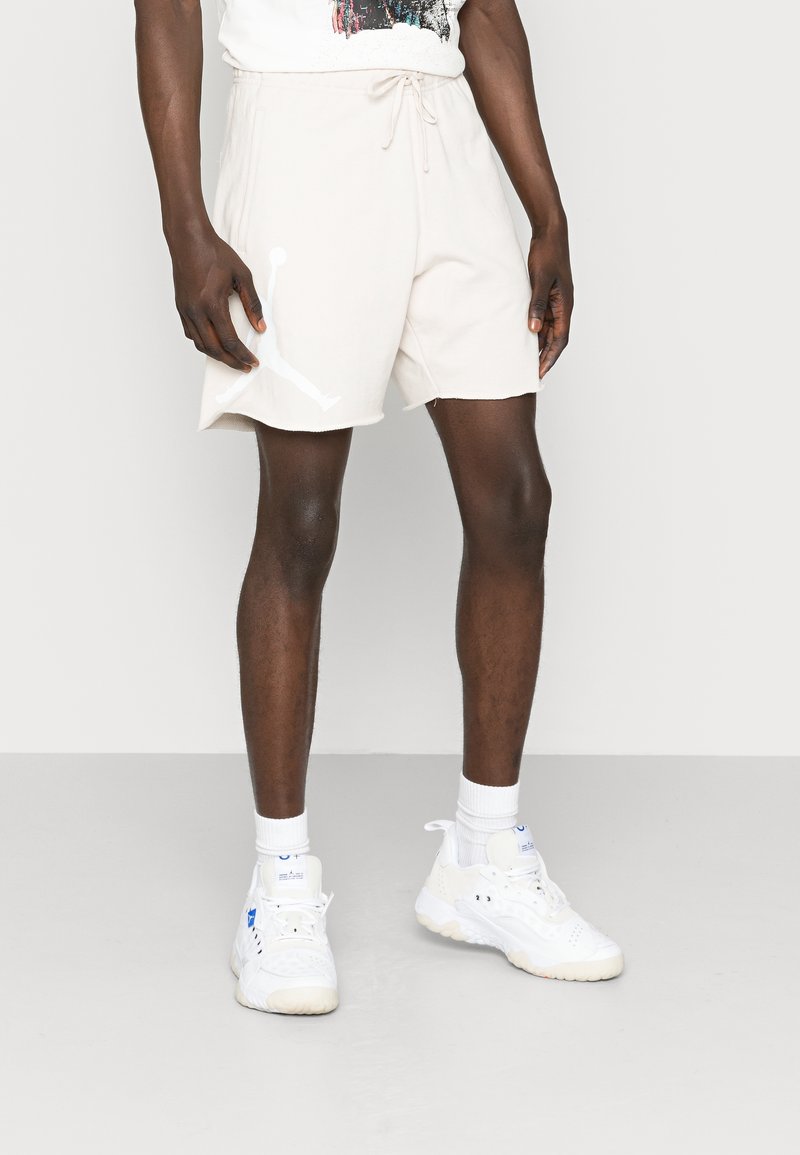 Shorts sportivi beige in cotone, con vita elasticizzata e cordino, e una grafica bianca del logo sulla gamba sinistra.