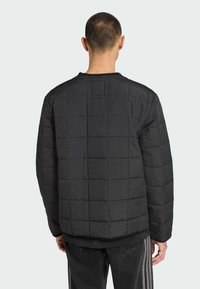 Een persoon staat met de rug naar ons toe, gekleed in een zwarte gewatteerde jas met een ruitpatroon, gecombineerd met donkere broeken met zijstrepen.