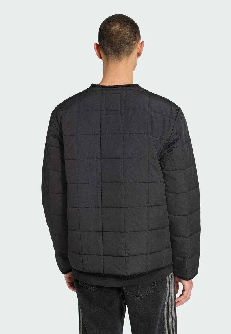 Une personne se tient de dos, portant une veste matelassée noire avec un motif en quadrillage, associée à un pantalon sombre avec des bandes latérales.