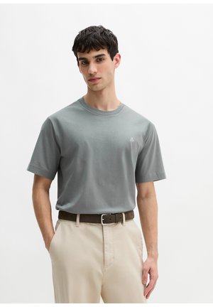 Junger Mann, der ein hellgraues kurzärmeliges T-Shirt und beige Hosen mit einem dunklen Gürtel trägt, vor einem schlichten weißen Hintergrund steht.