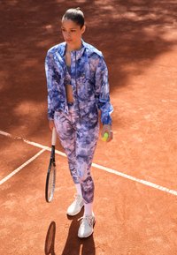 Modrá batikovaná sportovní bunda a legíny, bílé tenisky, držící tenisový míček a raketu, na antukovém hřišti s bílými čarami.