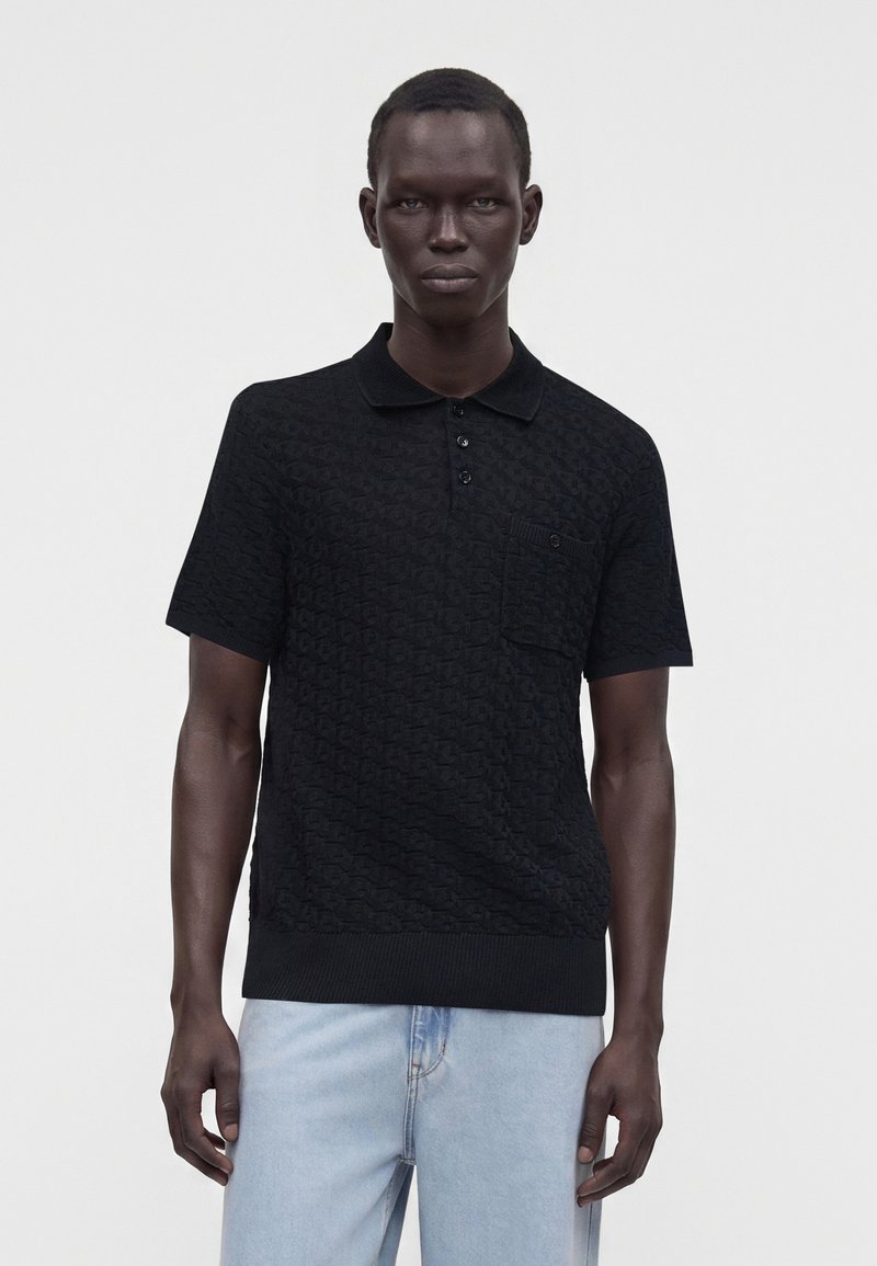 Homme portant un polo noir texturé à manches courtes avec des boutons et une poche poitrine, associé à un jean bleu clair, debout devant un fond uni.