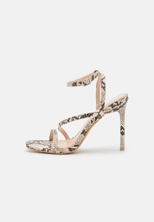 High-Heel-Sandale mit dünnen Riemen und Knöchelriemen in einem beigen und schwarzen Schlangenhautmuster vor einfachem Hintergrund.