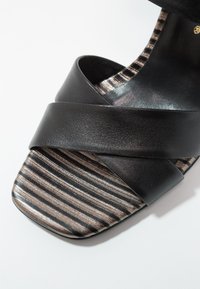 Sandale en cuir noir avec de larges brides croisées et un bout carré ornée de tissu métallique rayé aux tons noirs et bronze.