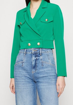 Kvinde iført en cropped grøn blazer med guldknapper kombineret med højtaljede lyseblå jeans på en lys baggrund.