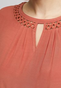 Blusa color corallo con scollatura rotonda, caratterizzata da ritagli circolari e un dettaglio a occhiello sul davanti. Realizzata in tessuto leggero e strutturato.