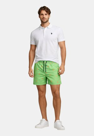 Mann, der ein weißes kurzärmeliges Poloshirt und grüne Badeshorts mit orangefarbenen Mustern trägt, steht mit weißen Sportschuhen vor einem schlichten Hintergrund.