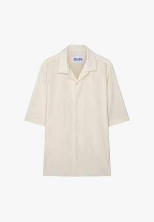 Ung mand iført en cremefarvet kortærmet skjorte med knapper, blå jeans og sorte pæne sko, stående foran en ensfarvet lys baggrund.