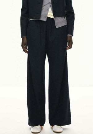Persona con pantalones negros anchos y sueltos, un cárdigan gris abotonado bajo una chaqueta negra corta y zapatos blancos sobre un fondo sencillo.