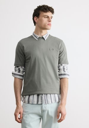 Młody mężczyzna patrzący w prawo, ubrany w warstwową, w paski koszulę na guziki pod szarym krótkim T-shirtem oraz jasnoniebieskie dżinsy.