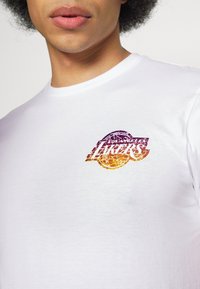 Kortärmad vit T-shirt med en flerfärgad grafisk logotyp som säger "Los Angeles Lakers" med en globdesign. Slät bomullstextur.