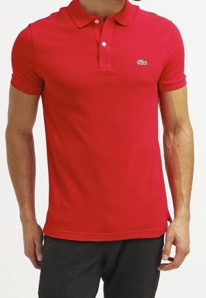 Mann trägt ein eng anliegendes rotes Poloshirt mit zwei weißen Knöpfen und kleinem grünem Logo auf der linken Brust, kombiniert mit schwarzer Hose.