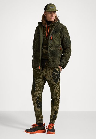 Chaqueta de forro polar verde con detalles en naranja, combinada con pantalones oscuros de flores verdes y zapatillas negras con detalles en naranja.