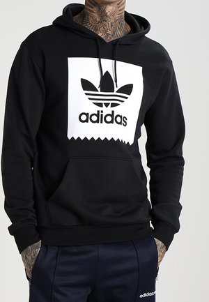 Osoba ubrana w czarną bluzę z kapturem Adidas z białym logo i granatowe spodnie Adidas, pokazująca tatuaże na szyi i rękach, stojąca na jednolitym tle.