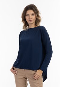 Mørkeblå langærmet top med løs pasform, rund halsudskæring og sideslids, lavet af glat stof. Bæres med beige bukser.