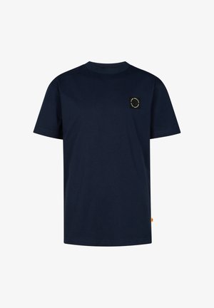Blond jongetje staand, draagt een marineblauw T-shirt, lichtgrijze wijde broek en witte sneakers tegen een effen lichte achtergrond.