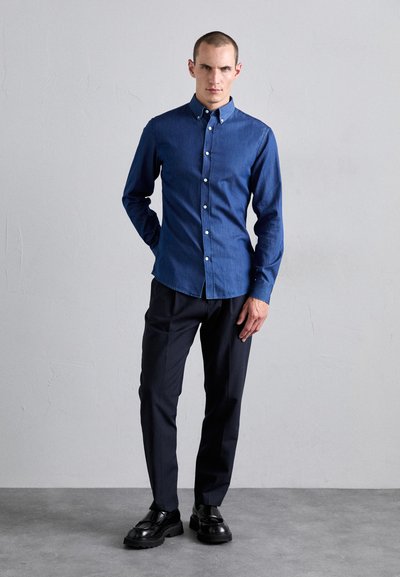 Cămașă din denim albastru, slim-fit, cu guler cu butoni, mâneci lungi, butoane albe și o textură netedă, purtată cu pantaloni închiși la culoare și încălțăminte neagră.