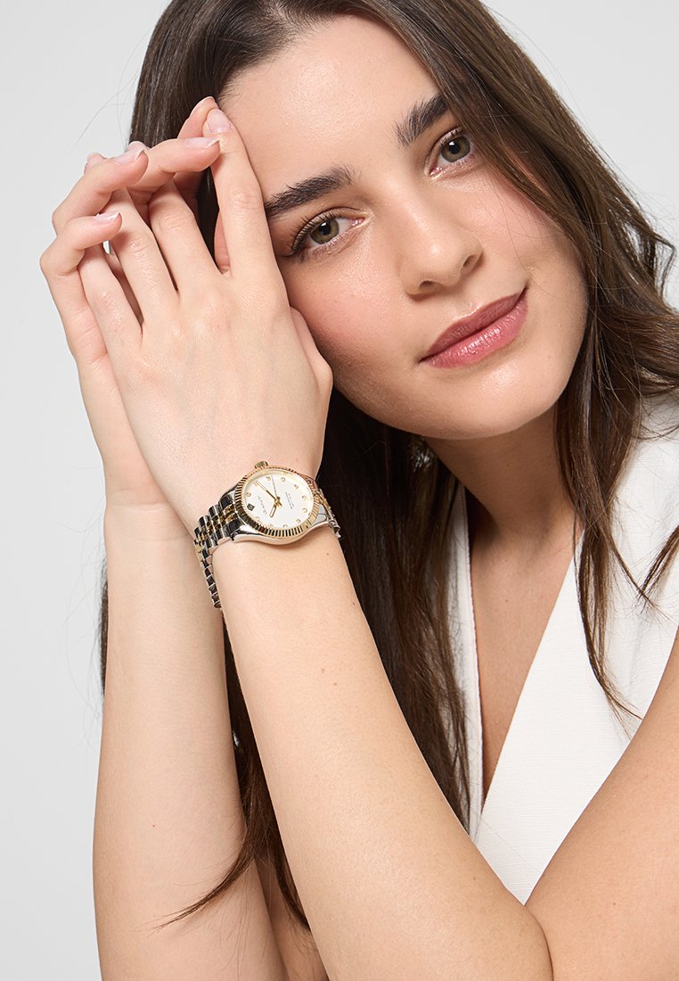 Montre en or et argent avec un cadran rond blanc, monté sur un bracelet à maillons. La main du modèle repose près de son visage, mettant en valeur la montre.
