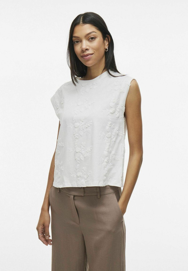 Top blanco sin mangas con bordado floral, escote redondo y corte relajado, combinado con pantalones de tiro alto marrón claro.