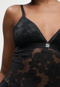 Γυναίκα που φοράει μαύρο φλοράλ δαντελένιο bralette με λεπτές τιράντες και ελαστική ζώνη κάτω από το στήθος με την ετικέτα "HUGO".