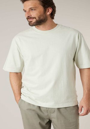 Man met baard, gekleed in een licht beige T-shirt met korte mouwen en kaki broek, staand met één hand in de zak, kijkt naar links.