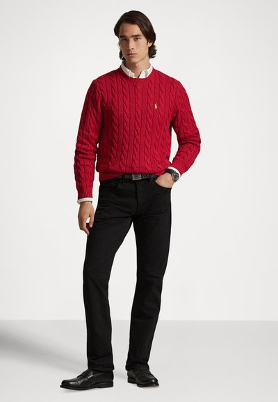 Άνδρας που φοράει κόκκινο πουλόβερ με πλέξη καλωδίου πάνω από λευκό πουκάμισο, μαύρο παντελόνι, μαύρη ζώνη, μαύρα παπούτσια και ρολόι χειρός, στέκεται μπροστά σε απλό φόντο.