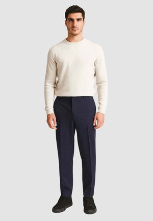 Maglione a maglia beige chiaro, pantaloni navy su misura e sneakers nere. Presenta un collo a giro e un design aderente, con texture lisce.