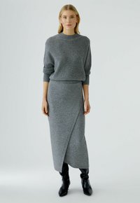 Pull en maille grise avec un col rond et une texture côtelée, associé à une jupe portefeuille grise. Des bottes noires à hauteur de genoux complètent la tenue.