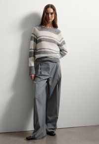 Strickpullover mit horizontalen grauen und weißen Streifen, Rundhalsausschnitt; kombiniert mit hochgeschnittenen, weit geschnittenen grauen Hosen, getragen mit schwarzen Schuhen.