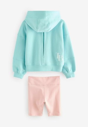 Vaaleansininen lyhyt huppari, jossa on etutasku ja TB-logo, yhdistettynä vaaleanpunaisiin pyöräilyshortseihin. Molemmat on valmistettu pehmeästä, joustavasta kankaasta.