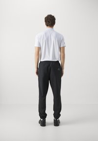 Black athletic pants with elastic cuffs and angled pockets, paired with a plain white short-sleeve shirt. Smooth fabric and fitted design.
