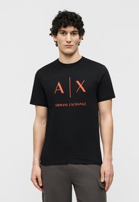Νεαρός με σγουρά μαλλιά που φοράει μαύρο T-shirt Armani Exchange με κόκκινο λογότυπο, στέκεται μπροστά σε απλό ανοιχτό γκρι φόντο.