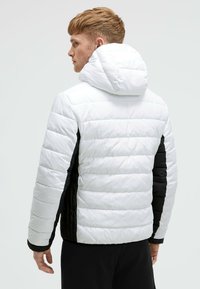 Chaqueta acolchada blanca y negra con capucha, con textura acolchada, cremallera frontal y paneles laterales. Ajuste entallado con puños elásticos.