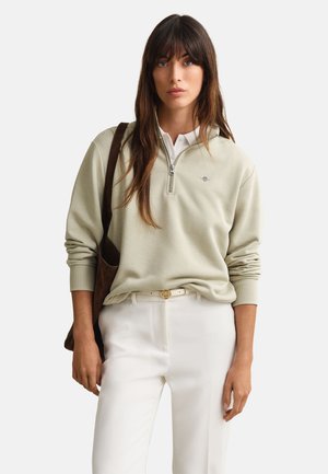 Žena s dugom smeđom kosom koja nosi bež džemper sa četvrtastim patentnim zatvaračem, bijele hlače, bijeli remen i nosi smeđu torbu preko ramena na jednobojnoj pozadini.