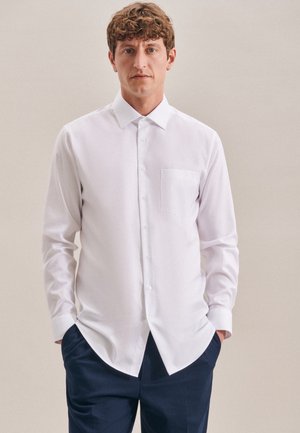 Camicia bianca a maniche lunghe con colletto a camicia, taschino singolo sul petto e una texture liscia, indossata con pantaloni blu scuro.
