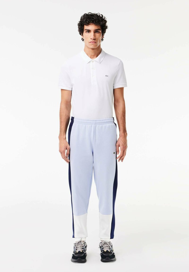 Polo blanc à manches courtes, associé à un jogging bleu clair avec des rayures latérales marine et des poignets blancs. Chaussures de sport noires.