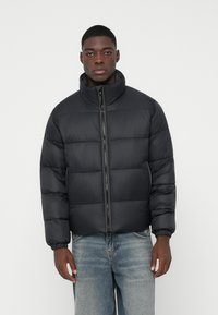 Schwarze Daunenjacke mit hohem Kragen, gesteppter Optik und Frontreißverschluss. Mit elastischen Bündchen und taillierter Silhouette, kombiniert mit blauen Jeans.