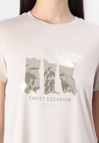 Kerge beež lühikeste varrukatega t-särk, millel on prinditud disain oliivrohelistest abstraktsetest kujunditest ja tekst "SWEET ESCAPISM" mustas kirjas.