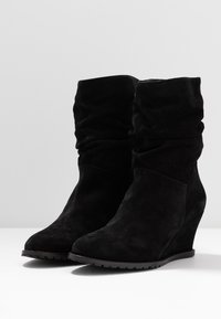 Bottes chevilles décontractées en daim noir avec talons compensés, présentant une texture lisse et un bout effilé. Design simple sans éléments métalliques visibles.