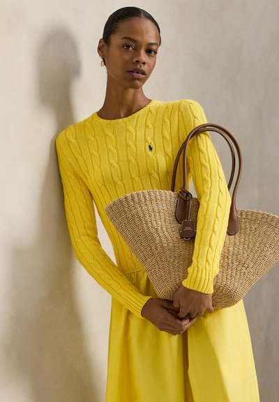 Κίτρινο πουλόβερ με πλέξη καλωδίου συνδυασμένο με αντίστοιχη φούστα. Το μοντέλο κρατά μια τσάντα ώμου από άχυρο με καφέ δερμάτινες λεπτομέρειες και ετικέτα.