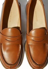 Mocasines de cuero marrón con textura lisa, detalles de costura y una correa decorativa en la parte superior. Logo de la marca visible en el interior.