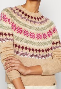 Osoba ubrana w beżowy sweter wełniany z maroonowymi, różowymi, białymi i zielonymi wzorami geometrycznymi oraz płatkami śniegu, z założonymi na piersiach rękami, nosząca dwa cienkie pierścionki.