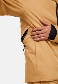 Giacca outdoor color sabbia dalla texture liscia, con tasca laterale con cerniera, dettagli neri e design aderente lungo il torso.
