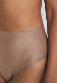 Bikini w wysokim stanie, bezszwowy, wykonany z jasnobrązowej tkaniny. Gładka struktura w minimalistycznym stylu bez widocznych elementów metalowych czy wzorów.