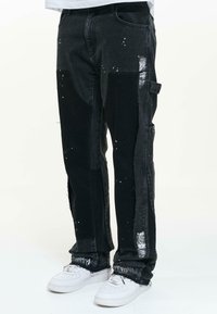 Mustat denim-housut, joissa on löysä malli, maalitahra-yksityiskohtia, kontrastiväriset kankaat ja sivutaskut. Tyylitetty valkoisilla lenkkareilla.